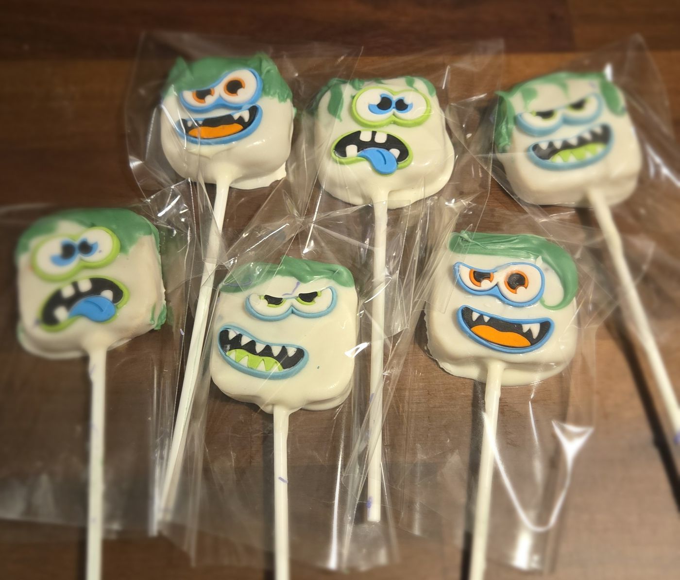 Six white chocolate monster cake pops with green hair and silly faces, wrapped in clear bags